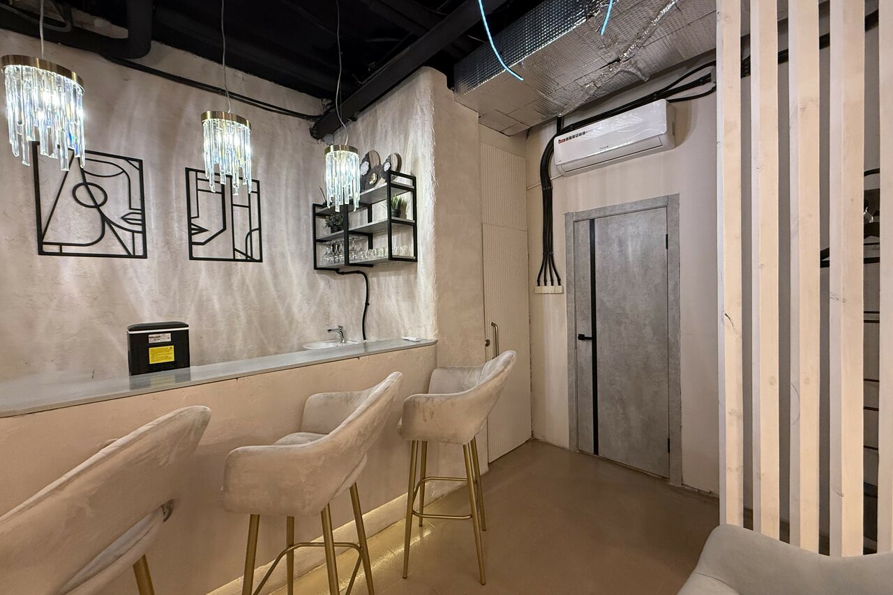 Арендовать Лофт SUNRISE(Восход Солнца). Просторное помещение на 15 гостей. Лофт-пространство с бетонными стенами и полом, барной стойкой с тремя бежевыми стульями. Подвесные светильники, декоративные элементы на стене, кондиционер, вертикальные деревянные панели. Дверь окрашена в серый цвет.
