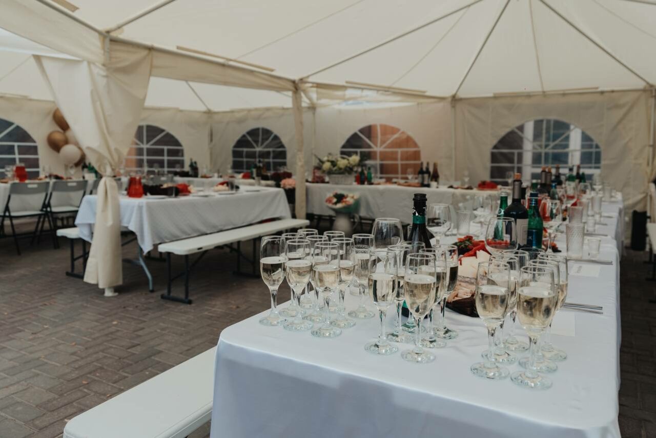 Арендовать Усадьба Афонинская. Белый шатёр с боковыми стенками из ткани и пластиковыми окнами. Внутри расставлены накрытые белыми скатертями столы с напитками и бокалами, деревянные лавки. Освещение естественное, пол выложен плиткой.