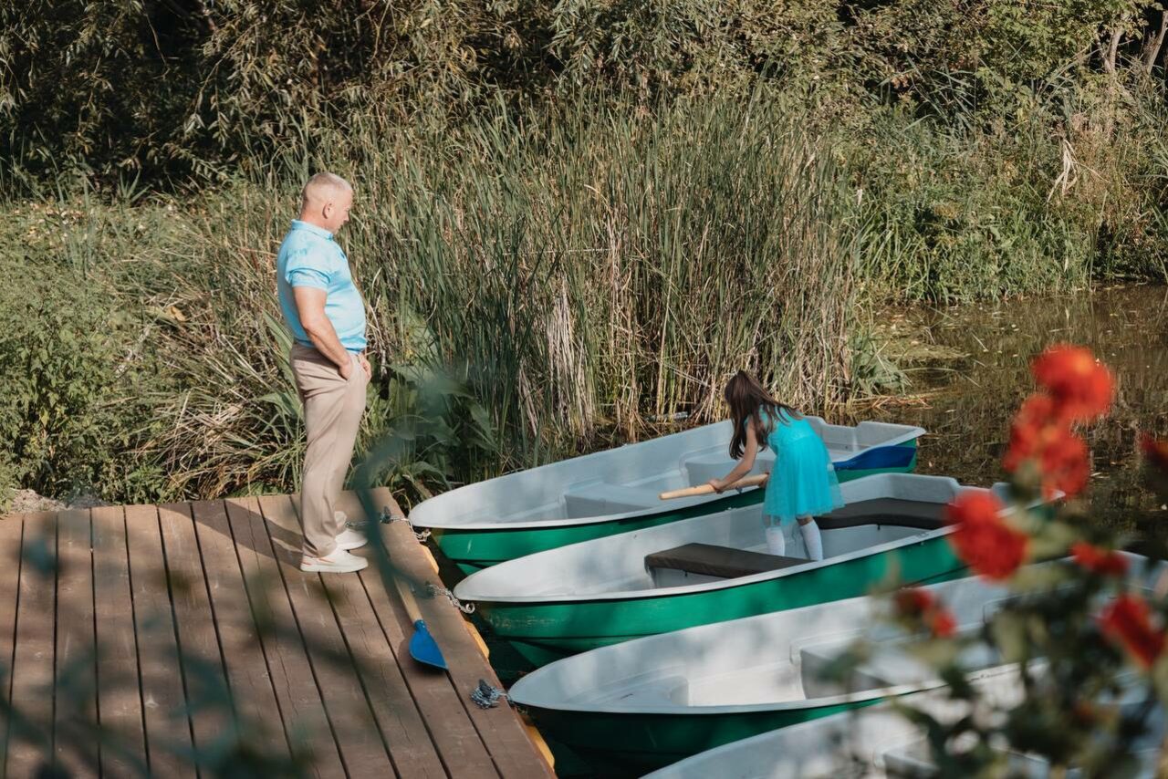 Арендовать Усадьба Афонинская. Деревянный причал у воды с тремя зелеными лодками, одна из которых занята ребенком. Взрослый стоит на причале. Вокруг густая растительность и цветы.