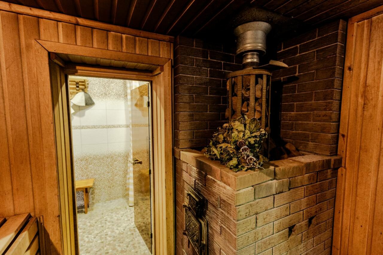Арендовать Усадьба Афонинская. Сауна с деревянной отделкой, справа кирпичная печь с камнями и металлическим дымоходом. Слева стеклянная дверь, за ней светлая комната с белыми плитками и стеной из мелких камней, деревянная скамья. Освещение тёплое.