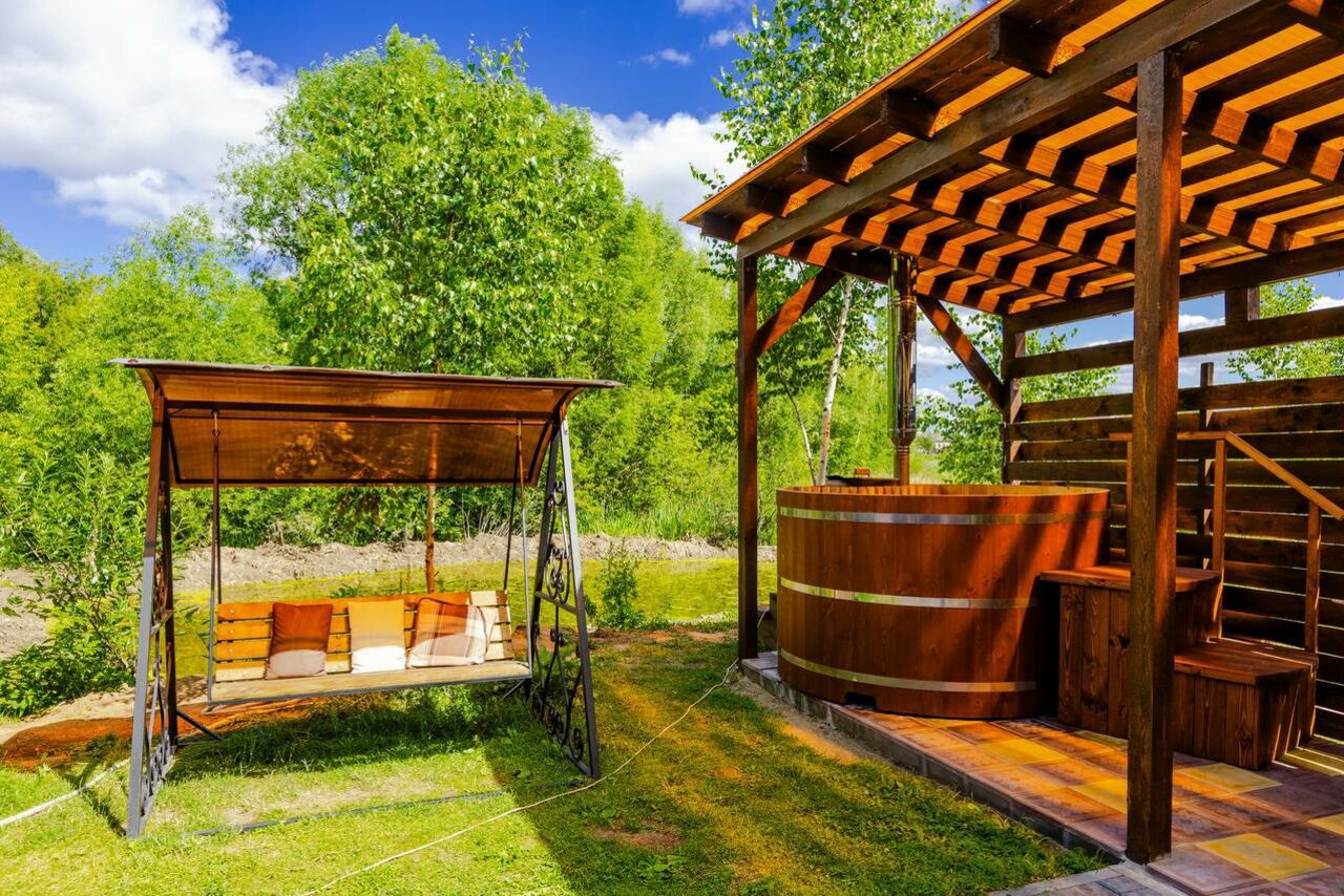 Арендовать Усадьба Афонинская. Открытое пространство с деревянной беседкой и деревянной купелью, справа деревянная терраса с перголой. Слева подвесные качели с подушками. Поверхность из травы и дерева, фон из деревьев. Яркое дневное освещение.
