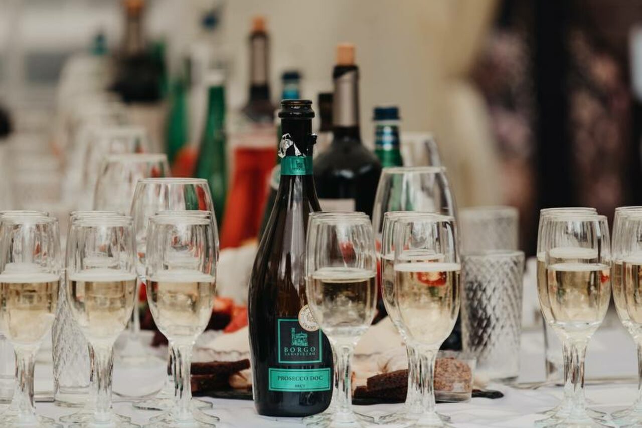 Арендовать Усадьба Афонинская. Стол с бокалами для вина и бутылками на фоне, в центре бутылка игристого вина с зелёной этикеткой, бокалы частично наполнены белым вином, вокруг другие стеклянные и зелёные бутылки, белая скатерть под ними.