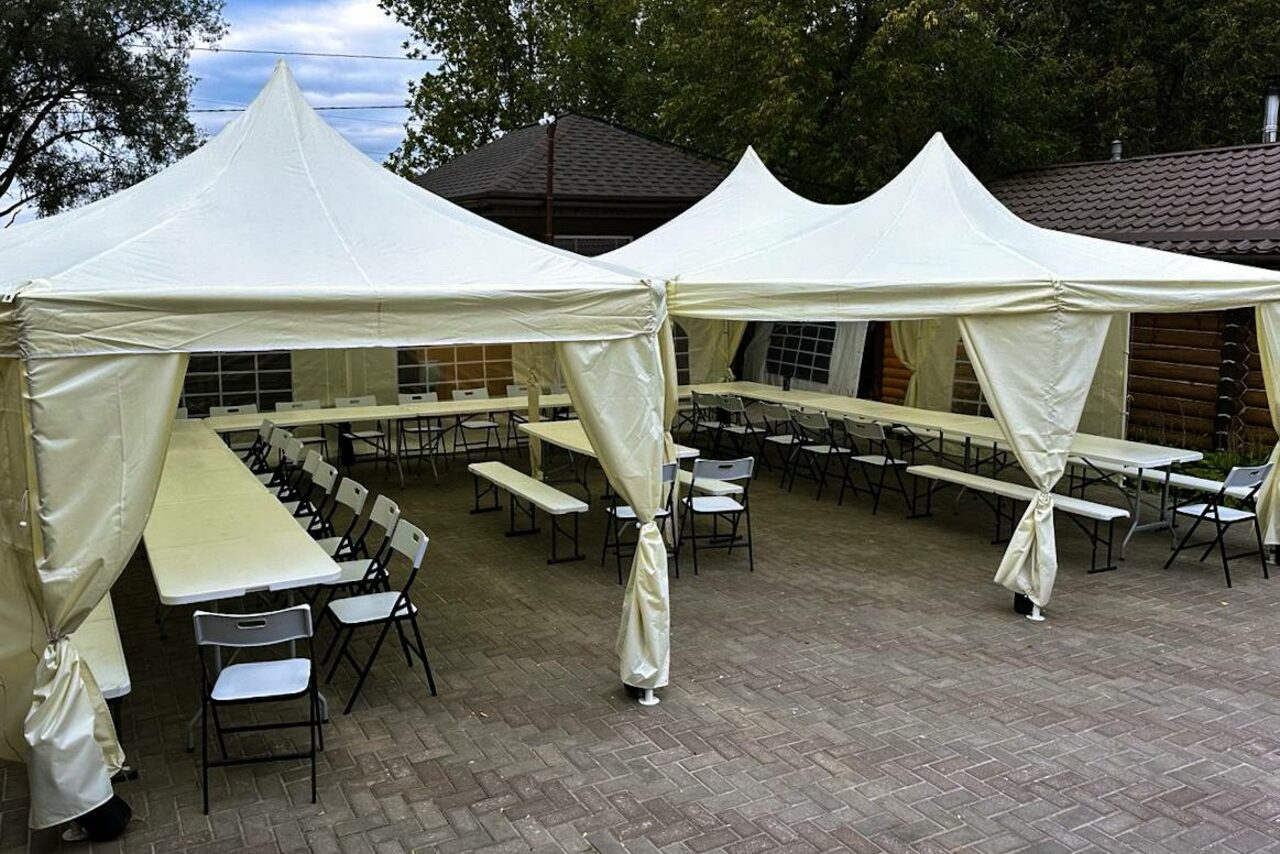 Арендовать Усадьба Афонинская. Площадка на открытом воздухе с двумя белыми шатрами, под которыми расставлены длинные столы и складные стулья. Мощеная брусчаткой территория. Шатры подвязаны, в глубине видна стена с окнами. Листья деревьев на заднем плане.