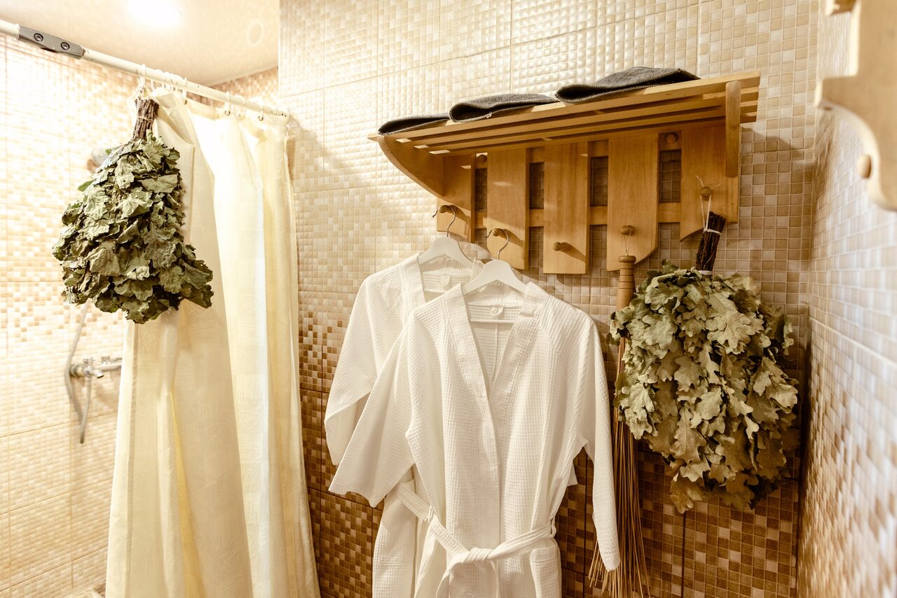 Арендовать Усадьба Афонинская. Сауна с плиточными стенами, вешалка с банными халатами и полотенцами, душевой уголок с занавеской, два веника на стене. Светлая отделка с деревянными элементами.