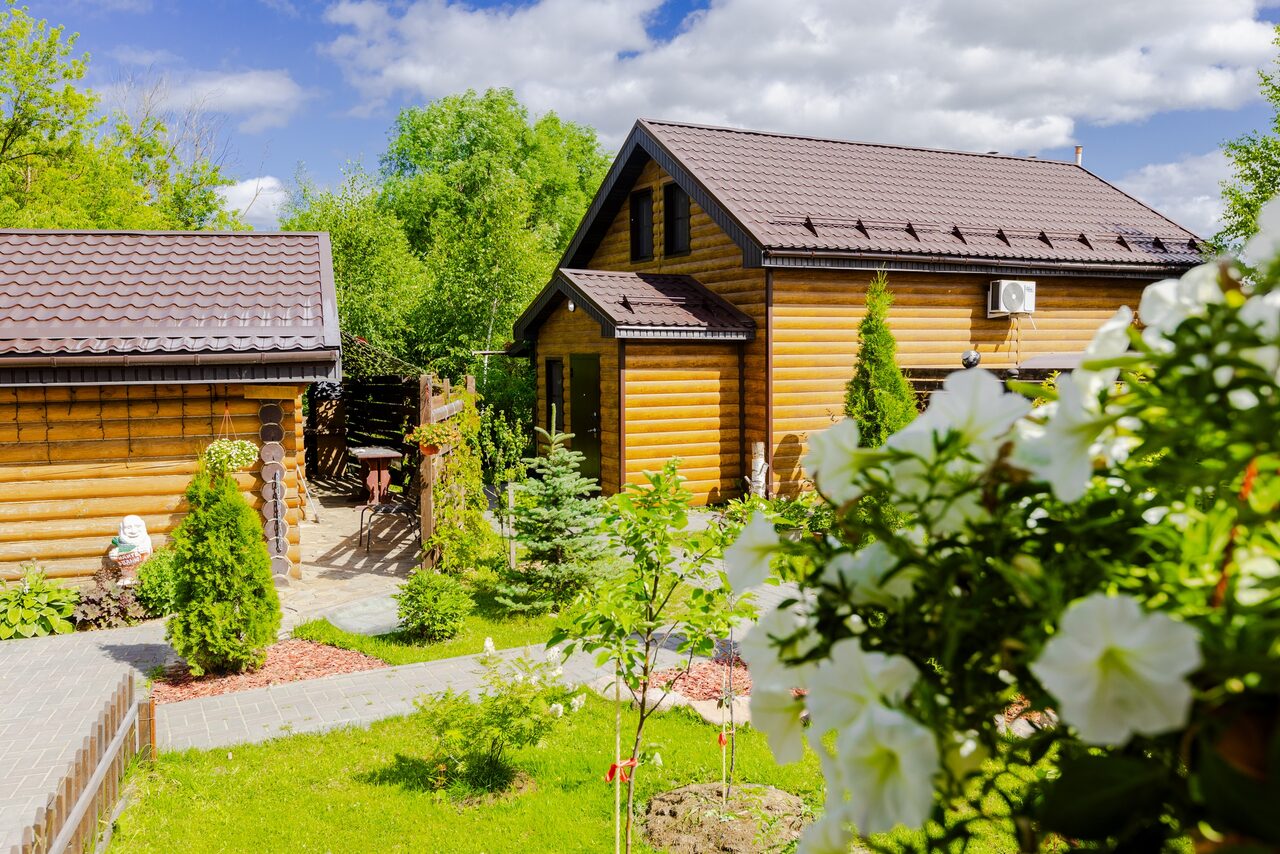 Арендовать Усадьба Афонинская. Два деревянных здания с коричневыми крышами, в окружении зелёного сада. Дорожка из кирпича ведет между домами. На переднем плане кусты с белыми цветами. Видна открытая беседка с садовой мебелью. Светлый солнечный день.