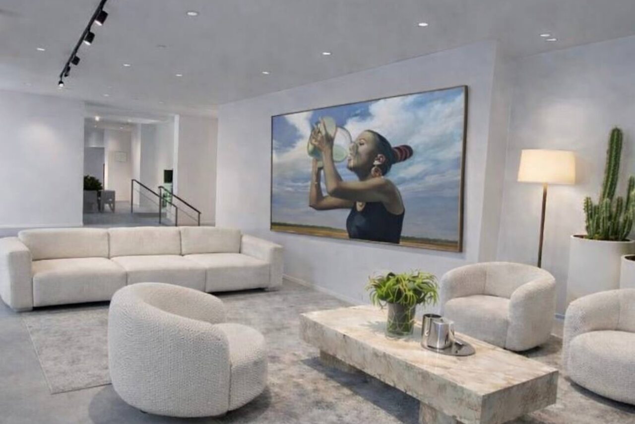 Арендовать Level 5(Лэвел 5). Современное пространство с мягким диваном и креслами белого цвета, центральный стол из светлого камня. На стене картина с изображением человека. Напольная лампа и кактус в углу. Светлый ковёр, потолочные светильники.