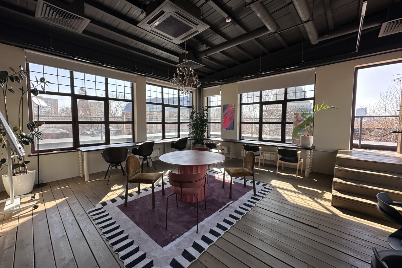 Арендовать Пространство с террасой до 50 человек. Просторная комната с деревянным полом, круглым столом и стульями. Большие окна с естественным светом, люстра, растения, ковёр с узором, картины на стенах. Черный потолок с трубами и кондиционерами, мягкие кресла у стен.