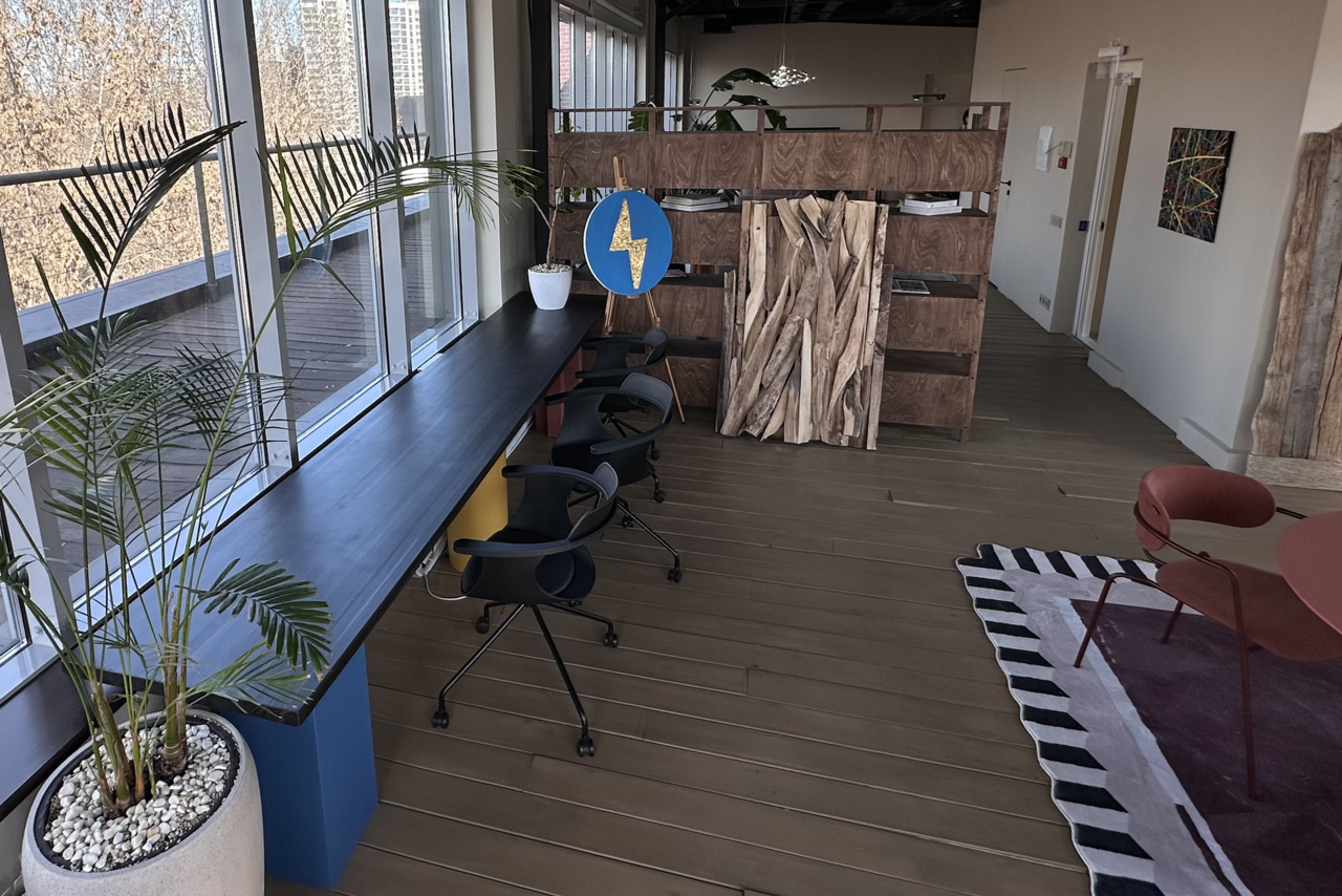 Арендовать Пространство с террасой до 50 человек. Современное офисное пространство с длинным столом, черными офисными креслами, деревянными перегородками. Большие окна, много естественного света. Плоские растения в горшках, деревянный пол, цветная мебель, декоративная зелень, абстрактная картина.