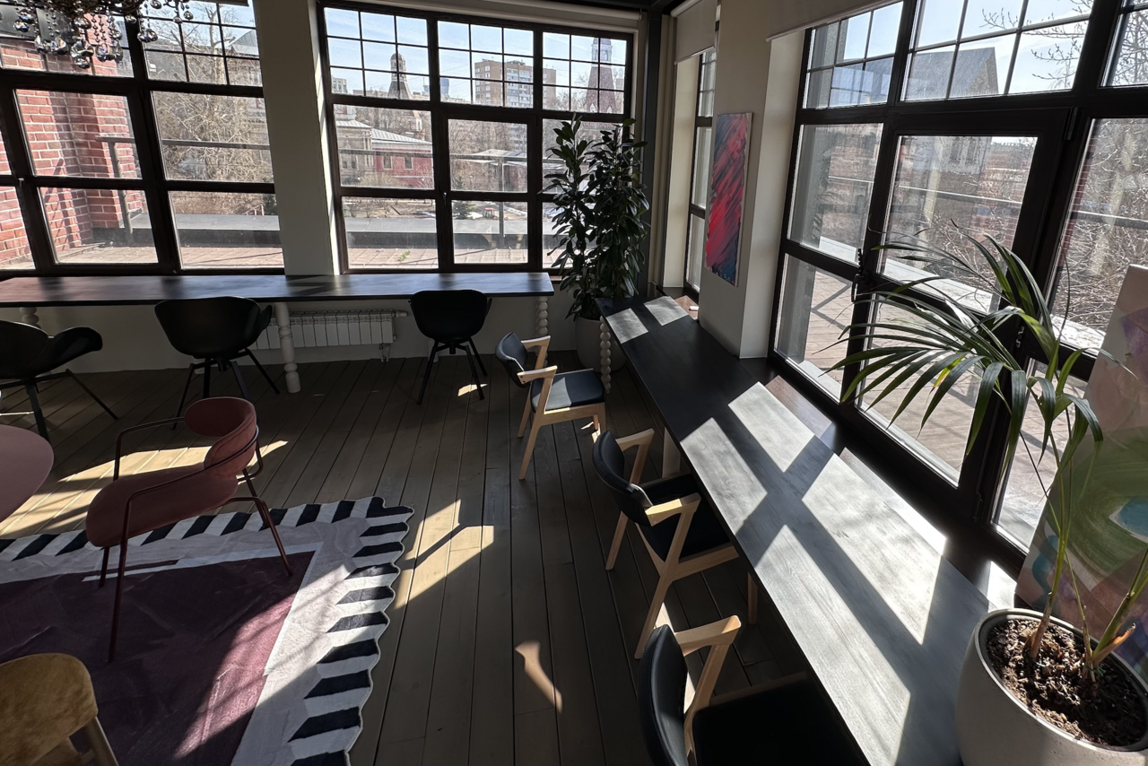 Арендовать Пространство с террасой до 50 человек. Лофт-пространство с деревянным полом и большими окнами. Вдоль окон столы с черными стульями. Растения в горшках. На полу ковер с геометрическим узором. Красно-кирпичная стена видна через окна.