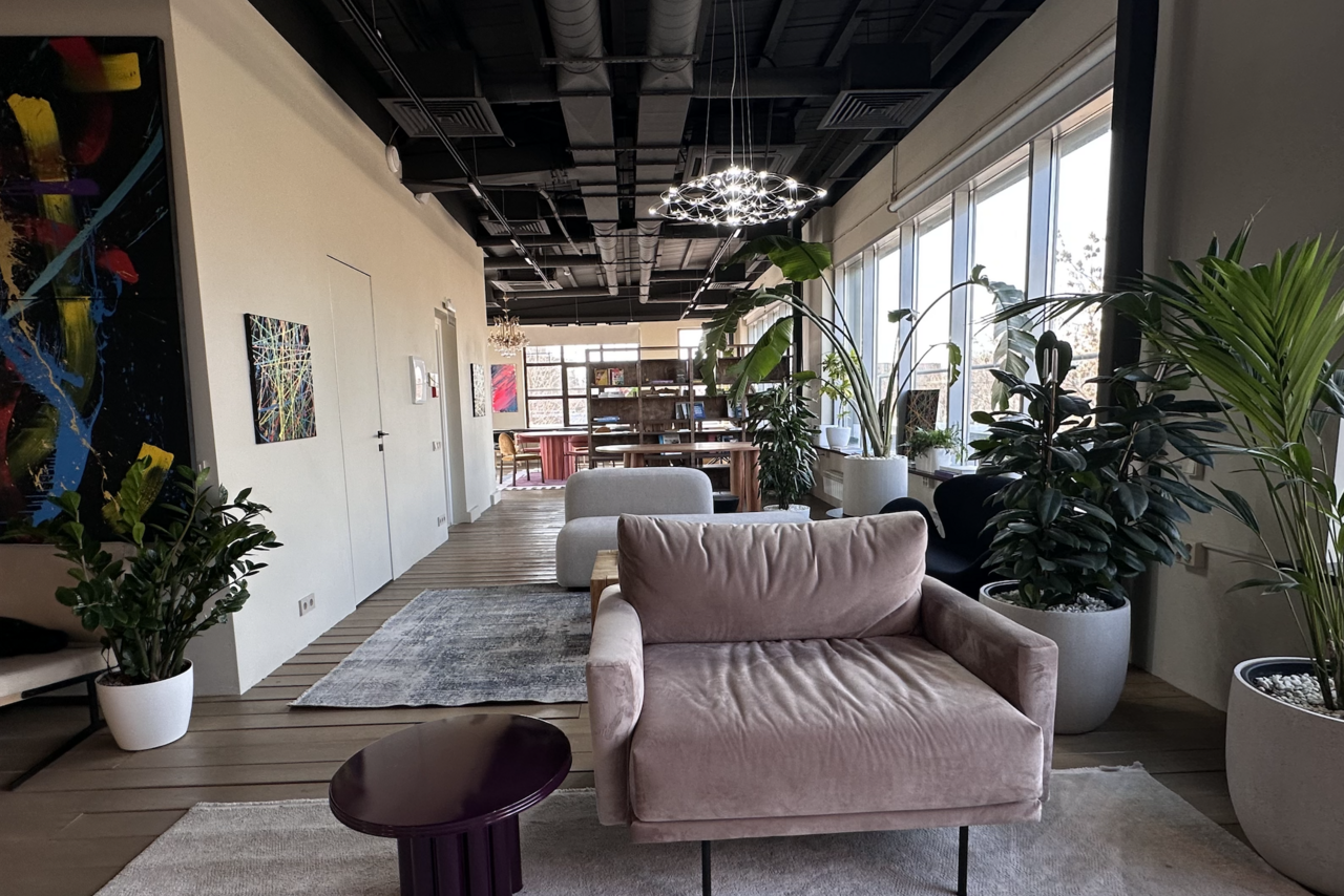 Арендовать Пространство с террасой до 50 человек. Лофт-пространство с окнами вдоль одной стены. Мягкий диван, журнальный столик и ковры на деревянном полу. Крупные горшечные растения. Декоративные картины на стенах. Подвесные светильники, белый и серый цвета, современный интерьер.