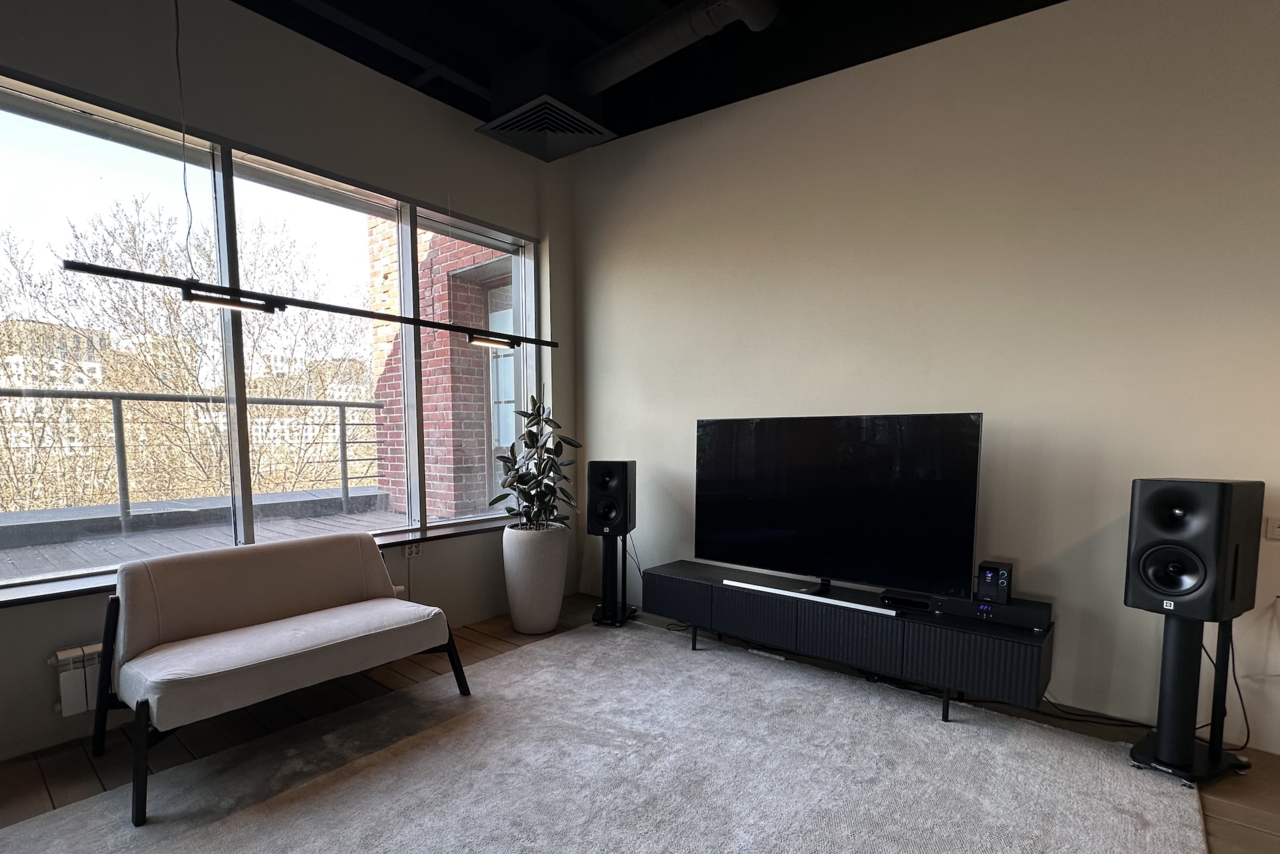 Арендовать Пространство с террасой до 50 человек. Минималистская гостиная с окном в пол. В комнате диван, телевизор на черной тумбе, колонки, ковер и пульт. Освещение подвесное, естественное светлое. Стены светлые, пол светлого дерева. Есть декоративное растение в белом горшке.