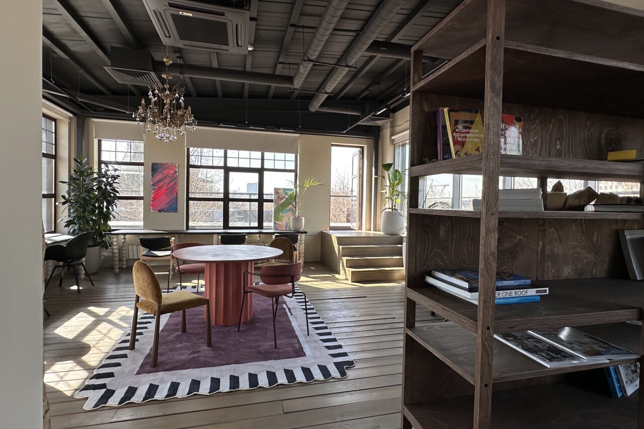 Арендовать Пространство с террасой до 50 человек. Просторное помещение в индустриальном стиле, деревянный пол, стеклянные окна, потолок с видимыми трубами. В центре круглый стол с разноцветными стульями на ковре, рядом стеллаж с книгами. На стенах картины, имеются растения и люстра.
