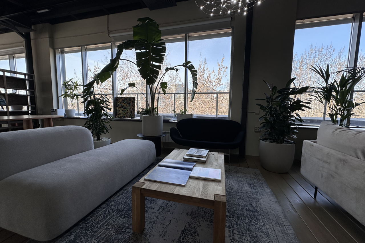 Арендовать Пространство с террасой до 50 человек. Просторная комната с большими окнами, современная мебель: светлый диван, темное кресло, деревянный стол. Декорирована растениями в горшках. На полу светлый ковер, потолок открытого типа. На столе журналы и книга.