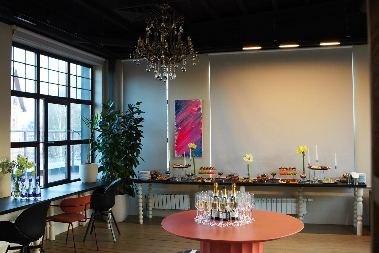 Арендовать Пространство с террасой до 50 человек. Пространство с большими окнами, круглым столом с напитками, стульями, барной стойкой, цветами, закусками. Светлые стены, деревянный пол. Потолочные светильники и люстра. Картина на стене.