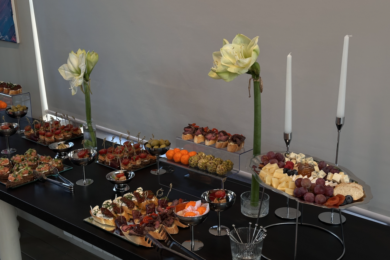 Арендовать Пространство с террасой до 50 человек. Стол с черной поверхностью, сервированный закусками, фруктами и сырами. Два высоких цветка и два белых свечи в подсвечниках. Стакан с вилками. Ровное освещение, светлые стены.
