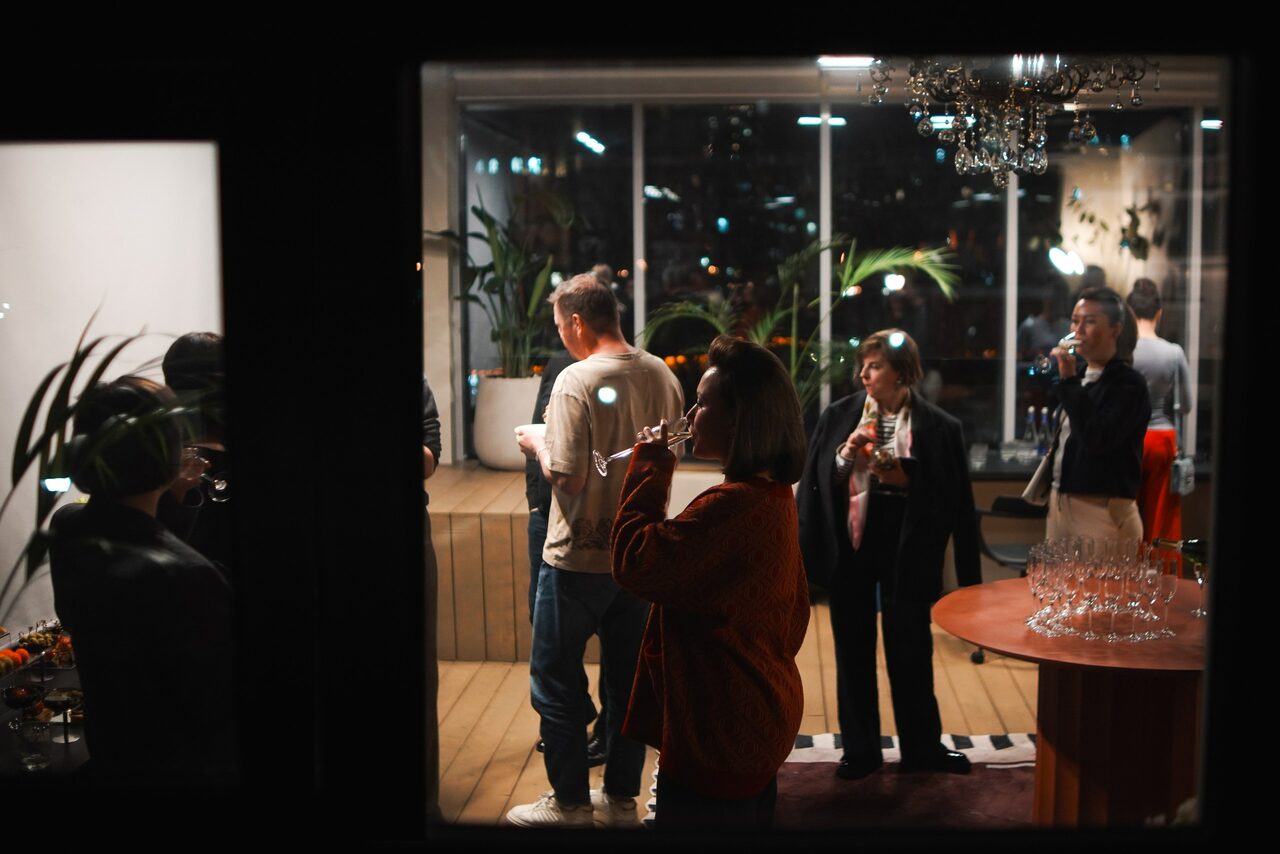 Арендовать Пространство с террасой до 50 человек. Современное интерьерное пространство с большими окнами, деревянным полом. Группа людей стоит у круглого стола с бокалами. В комнате растения, мягкое освещение, люстра. Ночной вид за окном.