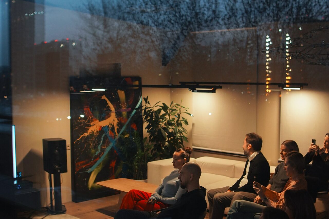 Арендовать Пространство с террасой до 50 человек. Современное помещение с большими окнами, белый диван, разноцветная картина на стене, зеленое растение, точечные светильники, деревянный пол. Четыре человека сидят, светится городской пейзаж за окном.