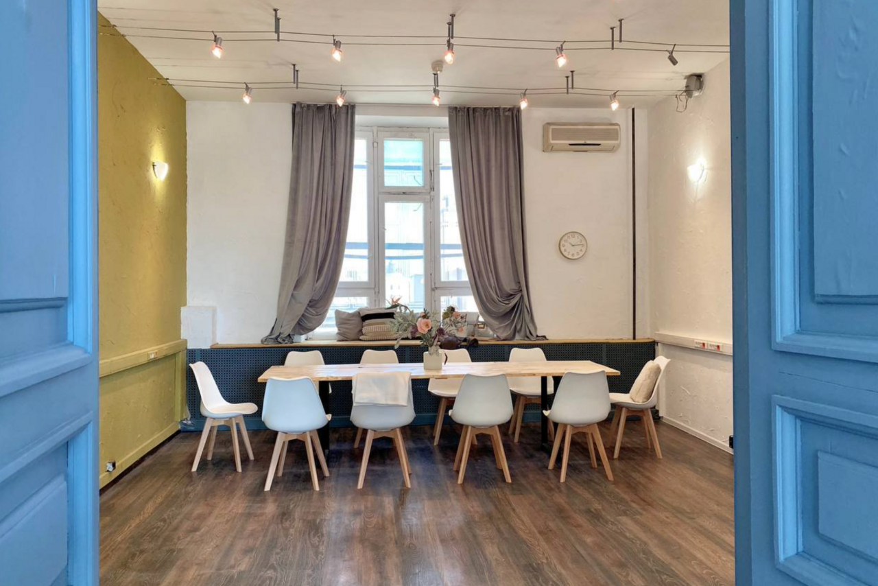 Арендовать H4U. Светлый зал П-602. Просторная комната с деревянным полом, большим светлым столом и белыми стульями. На стене часы, рядом окно с серыми шторами. Подвесные лампы на потолке, кондиционер на стене. Светлая и аккуратная атмосфера.