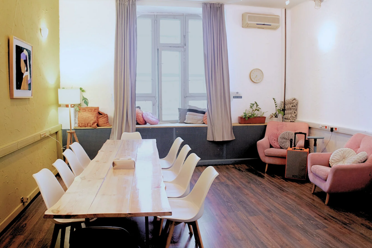 Арендовать H4U. Светлый зал П-602. Пространство с деревянным столом и стульями слева, розовыми креслами справа. На полу темное ламинатное покрытие, стены светлые. Окно с серыми шторами, лампа и растения. На стене часы, кондиционер, картина.