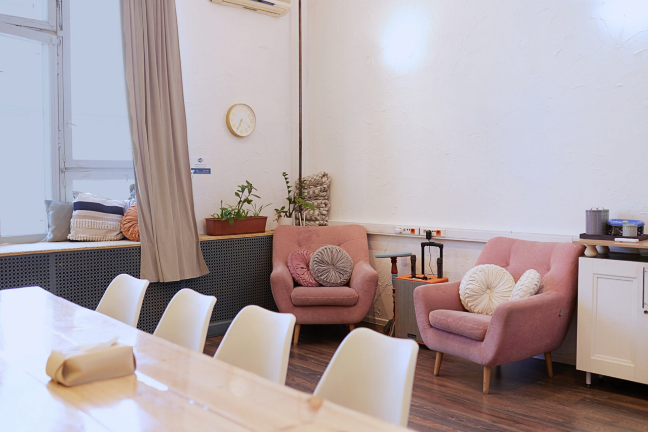 Арендовать H4U. Светлый зал П-602. Просторная комната с деревянным столом и белыми стульями. В углу находятся два розовых кресла с декоративными подушками и небольшой столик. На стене часы, на подоконнике подушки и растения. Светлое освещение, белые стены, деревянный пол.