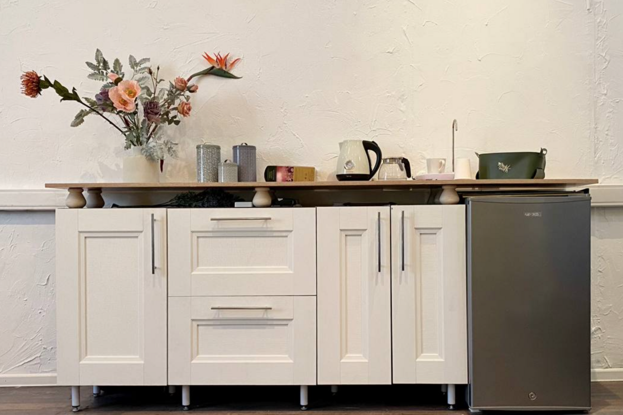 Арендовать H4U. Светлый зал П-602. Мини-кухня с белыми шкафами, черной металлической фурнитурой и темной столешницей. На столешнице стоят чайник, кружки, цветы и аксессуары. Справа черный холодильник. Белая текстурированная стена на фоне.