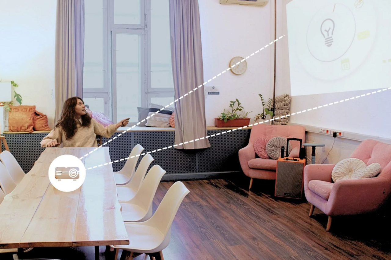 Арендовать H4U. Светлый зал П-602. Просторное помещение с деревянным столом и белыми стульями; на стене проекция изображения. В углу розовые кресла и подушки, между которыми небольшой столик и динамик. Пол тёмный деревянный. Есть окно с занавесками, комнатные растения и настенные часы.