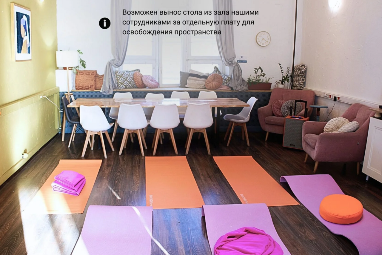 Арендовать H4U. Светлый зал П-602. Пространство с деревянным полом, длинным столом и белыми стульями. На полу фиолетовые и оранжевые коврики, подушки. Мягкие кресла, светлые стены. Свет через большое окно, есть растения и картина на стене. Текст на стене.
