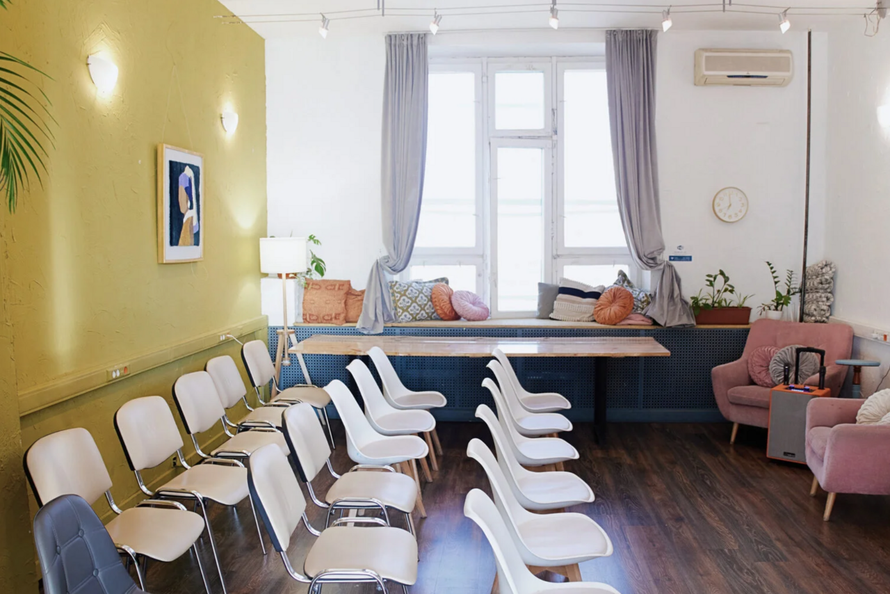 Арендовать H4U. Светлый зал П-602. Пространство с деревянным полом включает ряды белых стульев, два розовых кресла, подоконник с подушками, серые шторы и настенный кондиционер. Стены светлые с единичной ярко-зелёной секцией. На стене часы, картина и светильники.