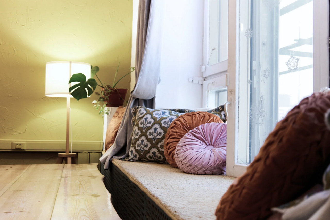 Арендовать H4U. Светлый зал П-602. Уютное пространство с окнами, деревянными полами и светлыми стенами. На подоконнике расположены декоративные подушки разных форм и узоров. В углу стоит торшер и растение в горшке. Светлая, спокойная атмосфера.