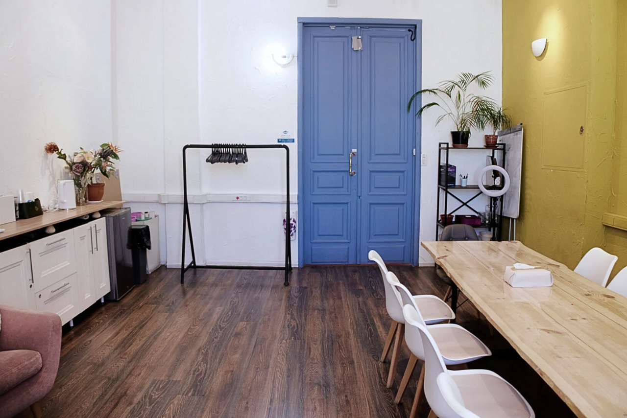 Арендовать H4U. Светлый зал П-602. Пространство с деревянным полом, синими двойными дверями, большим деревянным столом и белыми стульями. Слева комод с растениями и напитками. Справа стеллаж с лампой и зелёным растением. Белые стены с точечным освещением.