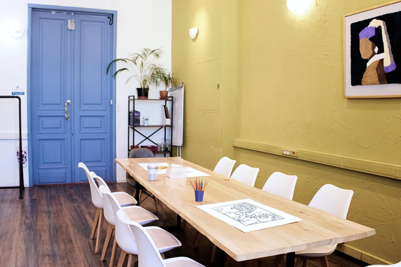 Арендовать H4U. Светлый зал П-602. Просторная комната с деревянным столом для собраний, окружённым белыми стульями. Слева дверь синего цвета, на стене зелёного оттенка светлое бра и картина. Пол тёмного дерева, на столе раскраска и цветные карандаши, растения на полке.