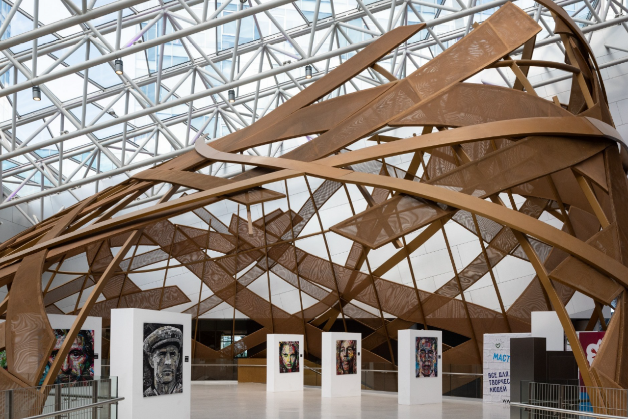 Арендовать Кунцево Плаза. Выставочное пространство с витиеватой структурой из коричневых деревянных элементов под стеклянной крышей. На белых стендах расположены портретные картины. Белый пол, металлические перила, знаки с текстами на заднем плане.