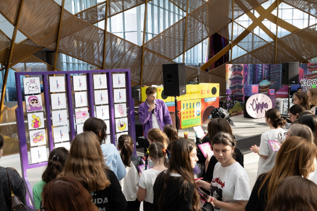 Арендовать Кунцево Плаза. Мероприятие в помещении с высокими окнами и геометрическими конструкциями на потолке. Множество людей смотрят на стенды с изображениями персонажей. Есть микрофон и колонки. Яркие цвета: фиолетовый, жёлтый, розовый.