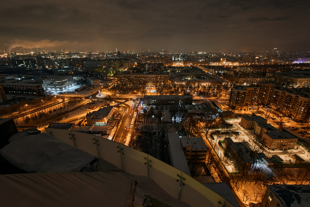 Арендовать 25 level (25 лвл). Лофт с панорамной крышей. Изображение ночного города с высоты, видны освещенные улицы, многоквартирные дома, деревья. На переднем плане частично виден обледенелый парапет. Освещение теплых тонов, небо темное с облаками.