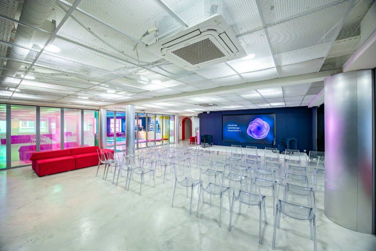 Арендовать АКИ.лаб пространство для мероприятий. Просторное современное помещение в стиле конференц-зала с серыми стенами и потолком из перфорированных панелей. Прозрачные стулья расположены рядами перед большим экраном на стене. Слева красный диван. Пол светлый, интерьер освещен ярким искусственным светом.