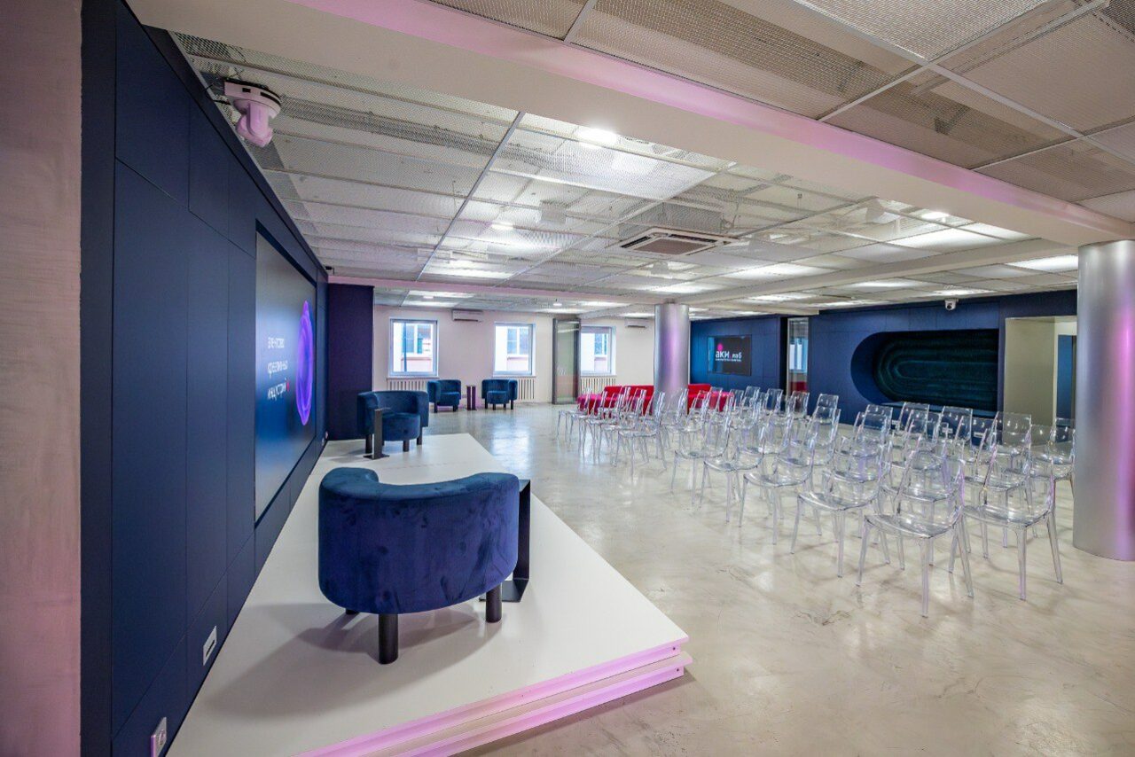Арендовать АКИ.лаб пространство для мероприятий. Современное конференц-пространство с панорамной планировкой и сценой, прозрачные стулья для зрителей, мягкие кресла на сцене. Большой экран, ровное освещение, бетонный пол, белый и синий цвета интерьера.