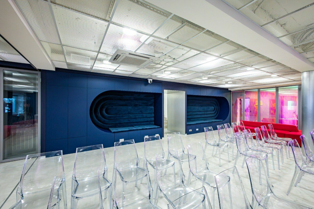 Арендовать АКИ.лаб пространство для мероприятий. Современное помещение со стеклянными стенами и прозрачными стульями. Синяя облицовка стен с нишами, белый потолок с подсветкой, кондиционер. Напольное покрытие светлое, вдоль стены красные диваны. Пространство предназначено для мероприятий.