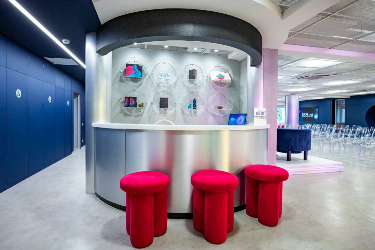 Арендовать АКИ.лаб пространство для мероприятий. Современное помещение с серебристой стойкой и тремя красными круглыми табуретами. На стене за стойкой круглые ниши с предметами. Пол бетонный, стены темно-синие. Освещение яркое, со встроенными светильниками.