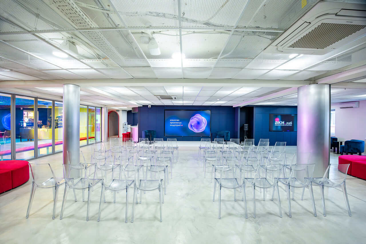 Арендовать АКИ.лаб пространство для мероприятий. Аудиторное пространство с прозрачными пластиковыми стульями, расположенными рядами. Светлые стены и потолок с встраиваемым освещением. Две металлические колонны. На стене мониторы с изображениями и текстом. Пол светлый, глянцевый.
