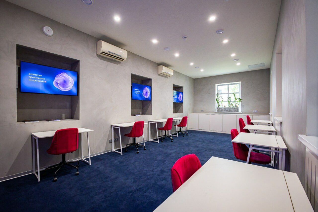 Арендовать АКИ.лаб пространство для мероприятий. Просторное помещение с серыми стенами и синим ковровым покрытием. Расставлены белые столы и красные кресла. На стенах установлены экраны. Под окном белая тумба. Потолочное освещение представлено светодиодными лампами.