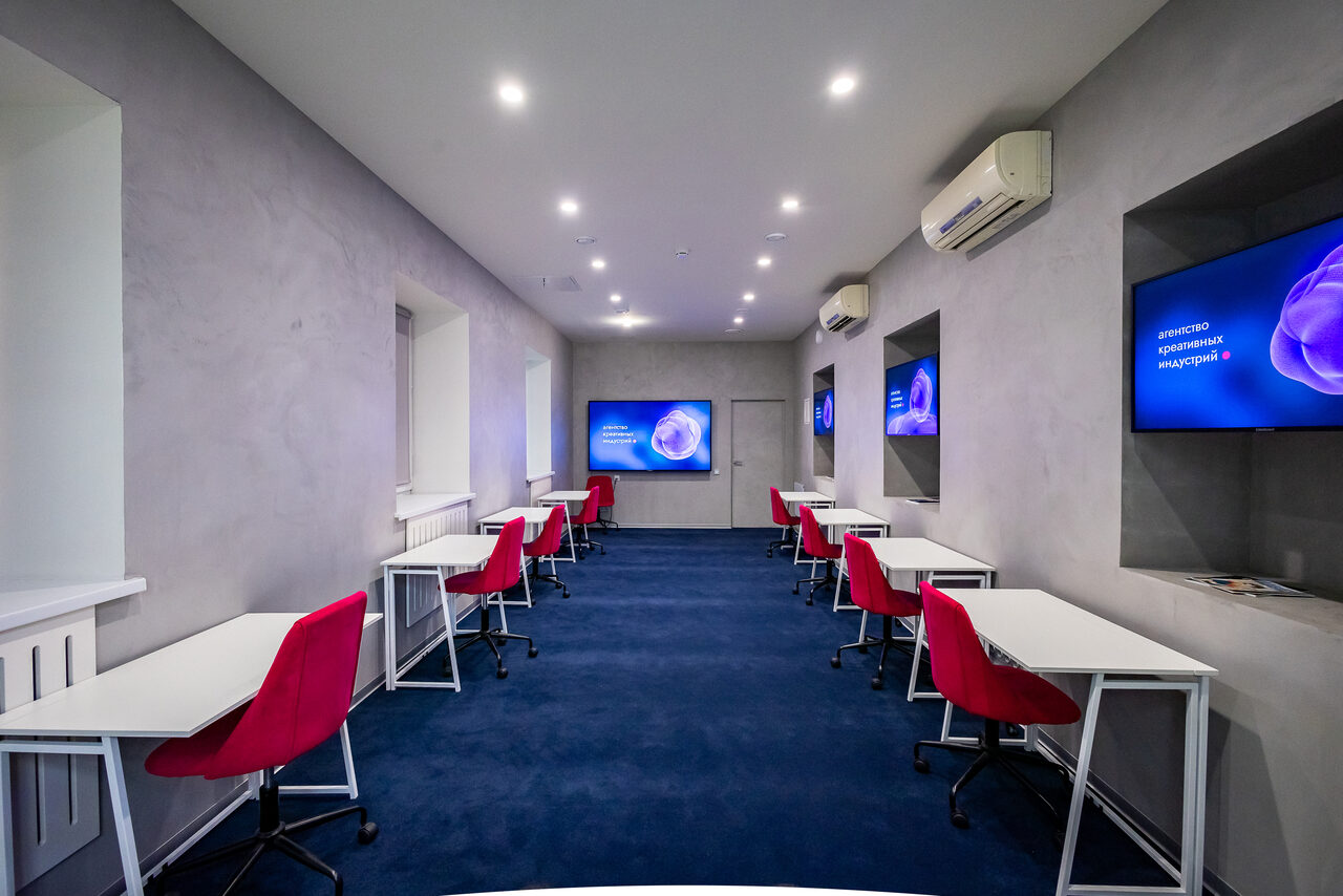 Арендовать АКИ.лаб пространство для мероприятий. Современное офисное помещение с рядами столов и красными стульями. На стенах крупные экраны, равномерное освещение потолочными светильниками, пол сине-зеленый, стены серые. Установлен кондиционер, окна обеспечивают естественный свет.