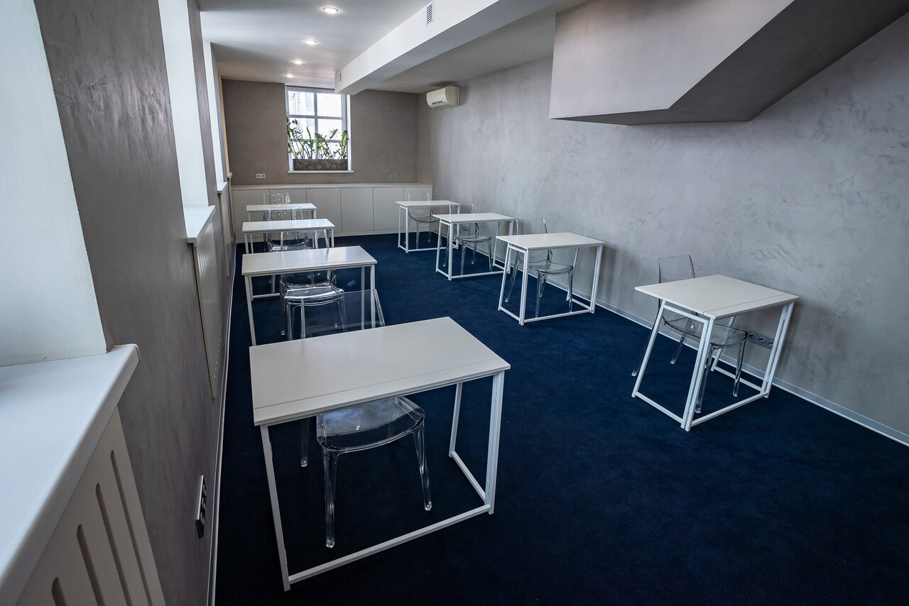 Арендовать АКИ.лаб пространство для мероприятий. Учебное помещение с несколькими белыми столами и прозрачными стульями на синем ковровом покрытии. Серые стены, потолочные светильники, окно с растением на подоконнике и кондиционер на стене.