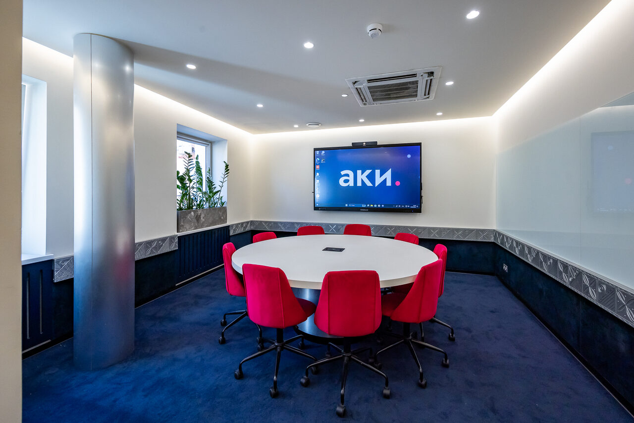 Арендовать АКИ.лаб пространство для мероприятий. Современная переговорная комната с круглым столом и восемью красными креслами. На стене крупный экран с текстом. Окно с растениями, колонна, белые стены, декоративные панели, встроенное освещение, система кондиционирования, синий ковролин.