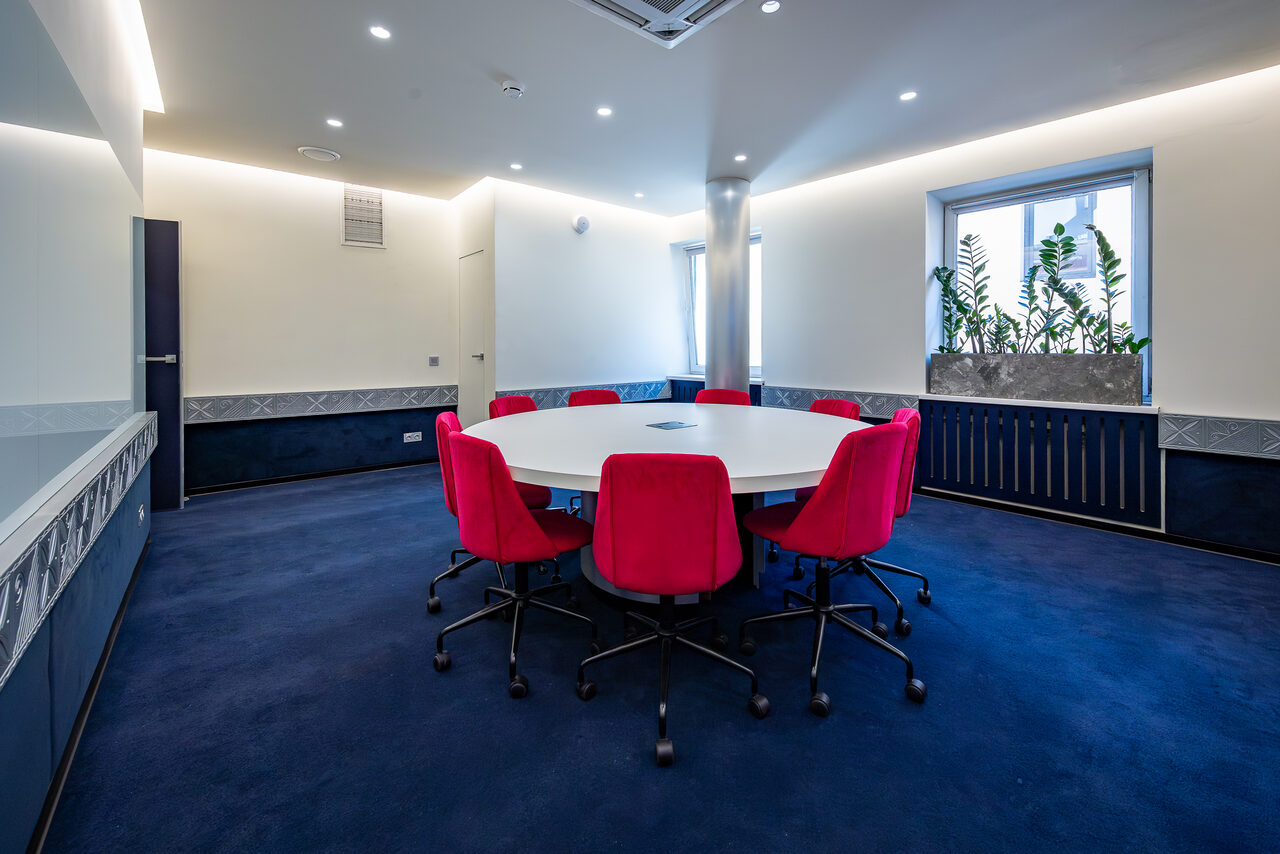 Арендовать АКИ.лаб пространство для мероприятий. Современное конференц-пространство с круглым столом и ярко-красными стульями на колесиках, синее ковровое покрытие, светлые стены с темными панелями. Окно с растениями, встроенное потолочное освещение и вентиляция.
