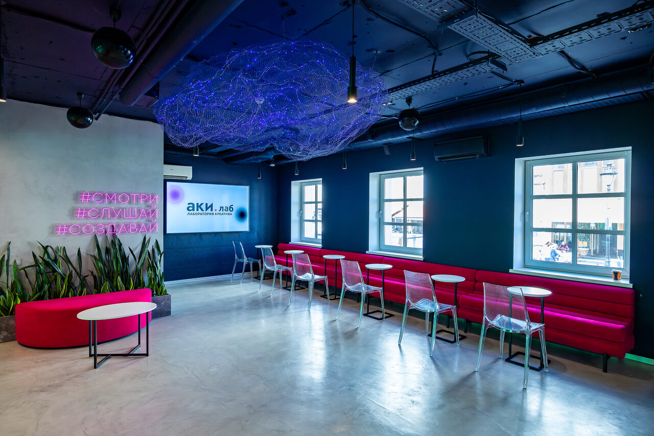Арендовать АКИ.лаб пространство для мероприятий. Пространство с бетонным полом, темным потолком и синими стенами. Есть красные диваны вдоль стены, прозрачные стулья, круглые белые столы. На стене неоновая надпись, в центре свисающая арт-инсталляция. Натуральное освещение из окон.