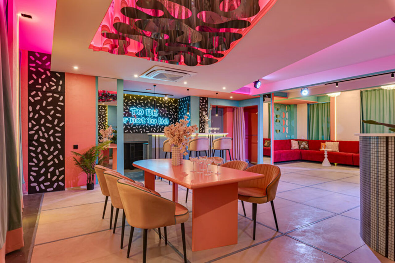 Арендовать Лофт Шекспир. Стильный дизайнерский зал. Современное пространство с открытой планировкой. В центре розовый стол и стулья, барная стойка с высокими стульями, разноцветные мягкие диваны, неоновая вывеска. Светодиодное освещение, разноцветные стены и декор, пол из светлых плит.