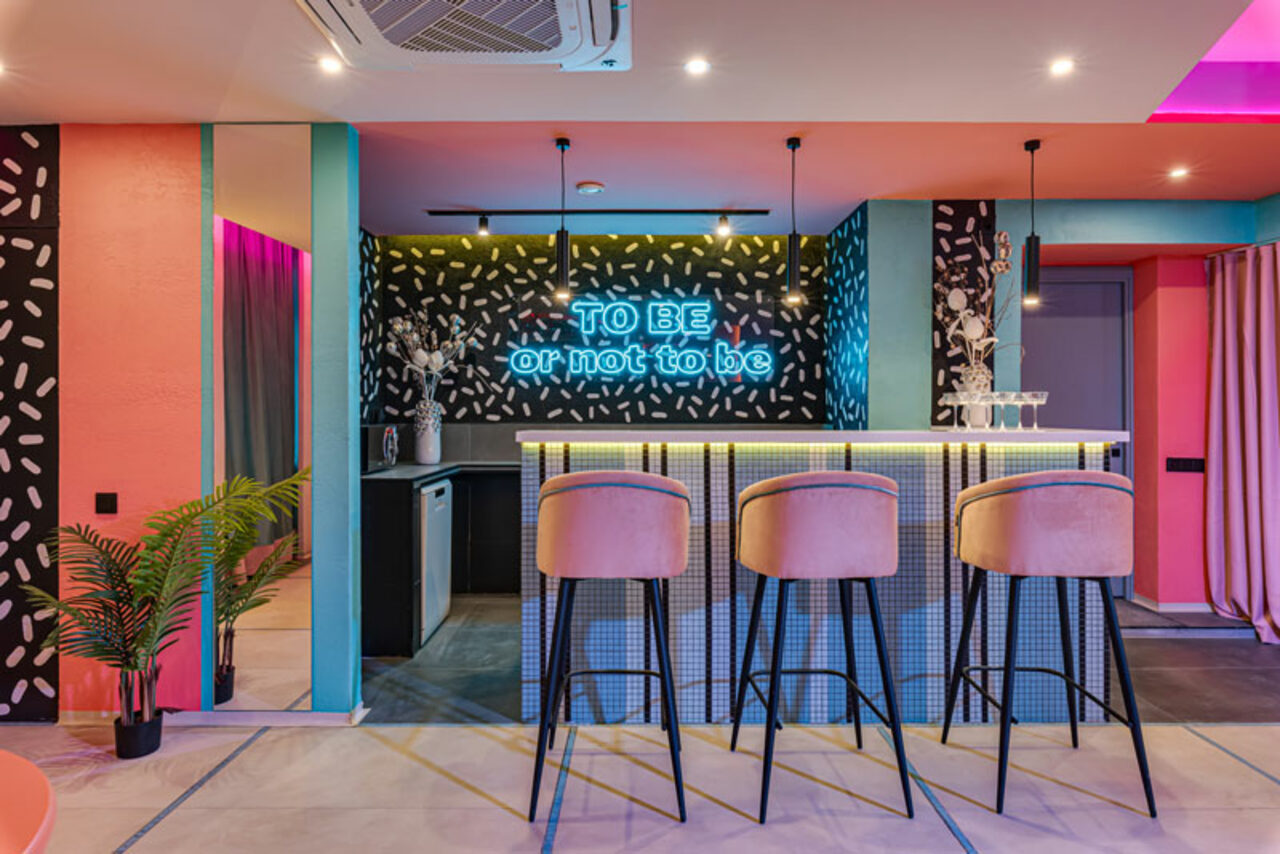 Арендовать Лофт Шекспир. Стильный дизайнерский зал. Барная зона с высокой стойкой и четырьмя барными стульями. На фоне неоновая надпись "TO BE or not to be". Стены декорированы черно-белыми узорами. Освещение подвесное, теплое. Видно растение в горшке и занавеску.