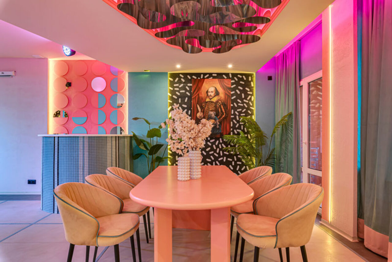 Арендовать Лофт Шекспир. Стильный дизайнерский зал. Пространство с длинным розовым столом и бежевыми стульями, декор с цветами на столе. Яркое освещение с розово-фиолетовыми неоновыми акцентами. На стенах постер с портретом, искусственные растения, шторы и барная стойка с круглым узором.