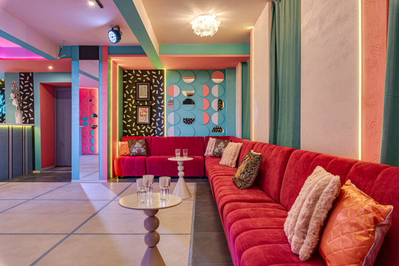 Арендовать Лофт Шекспир. Стильный дизайнерский зал. Просторная комната с длинным красным диваном вдоль стены, разноцветными декоративными панелями на стене, круглыми столиками с прозрачными стаканами, мягкими подушками. Яркое освещение, плиточный пол, разноцветные стены в геометрическом стиле.