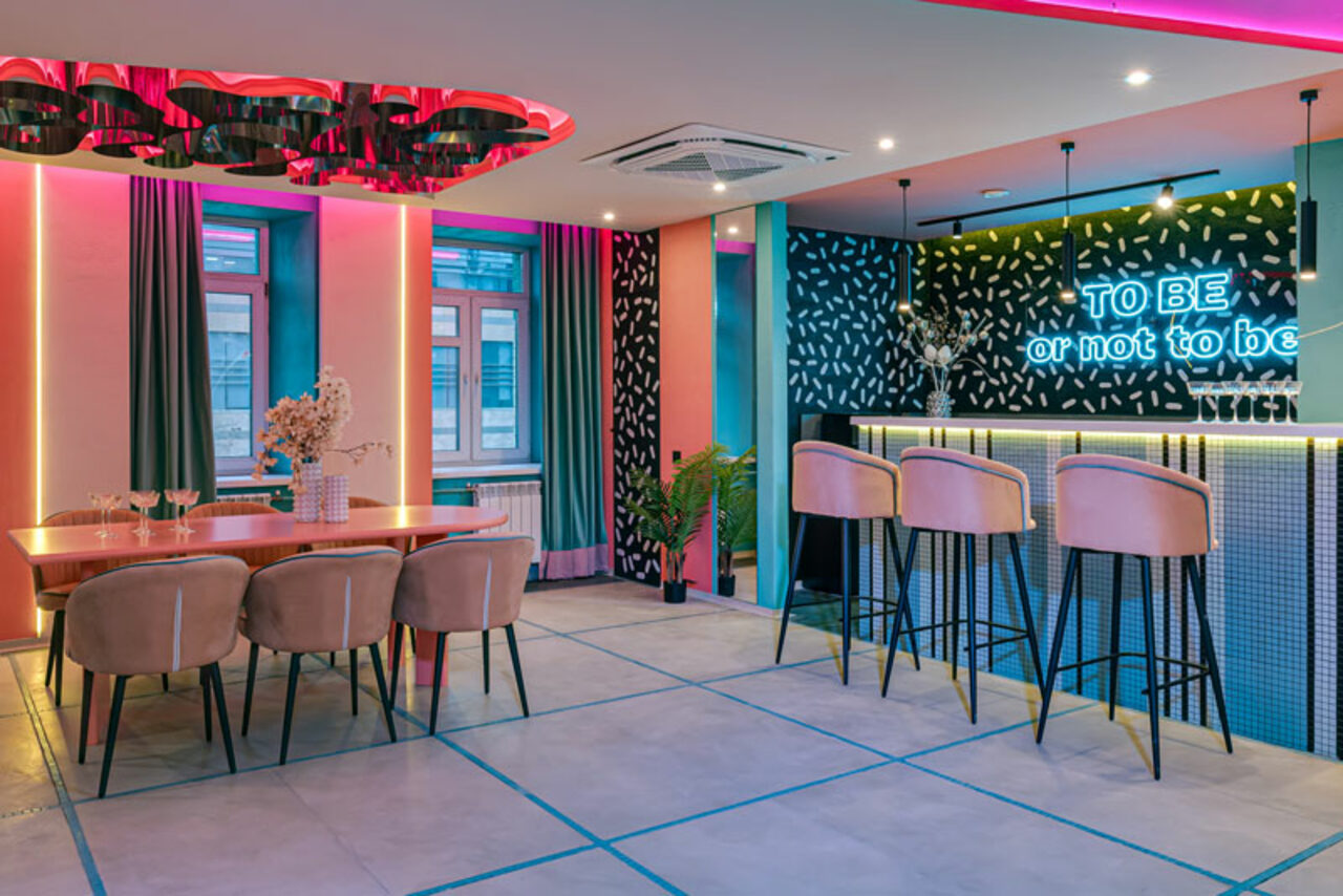 Арендовать Лофт Шекспир. Стильный дизайнерский зал. Современное пространство с обеденной зоной и барной стойкой. Зал украшен неоновыми надписями, имеет яркую цветовую подсветку. Мебель включает длинный стол с креслами и барные стулья. На стенах контрастные узоры. Пол из светлой плитки.